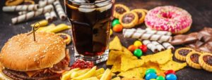 Varietà di cibo spazzatura con hamburger, patatine fritte, soda e dolciumi, con la scritta "CIBO SPAZZATURA E ADOLESCENTI" e il logo di Scienze Motorie