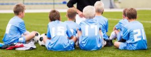 Bambini con maglie azzurre numerate seduti su un campo da calcio mentre ascoltano un allenatore; un pallone è accanto a ciascuno.