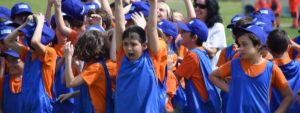 Bambini con magliette arancioni e gilet blu che partecipano con entusiasmo a un’attività sportiva all’aperto.