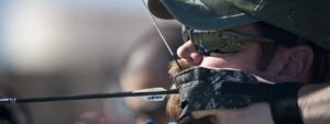 Persona con cappello, occhiali da sole e guanti da tiro che tende un arco con una freccia, mirando con concentrazione verso il bersaglio.