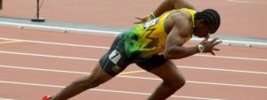 Atleta in posizione di partenza su pista d’atletica con divisa gialla, verde e nera, scarpe rosse e il testo “Efficienza e potenza nella corsa” sotto l’immagine.