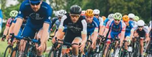 Gruppo di ciclisti con caschi e abbigliamento sportivo aderente colorato, concentrati in gara su strada all’aperto con alberi sullo sfondo.