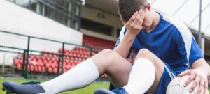 Calciatore seduto a terra con la testa tra le mani e un pallone accanto, su un campo da gioco con tribune vuote.