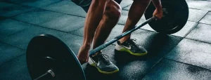 Parte inferiore del corpo di persona che solleva un bilanciere in palestra con scarpe sportive e piastrelle di gomma
