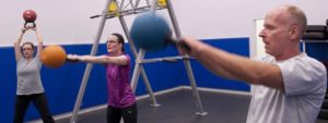 Tre persone che sollevano kettlebell all’altezza del petto in una palestra spaziosa con pareti blu.