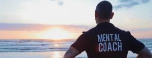 
L'immagine mostra la schiena di un uomo, presumibilmente un adulto, in piedi su una spiaggia, che osserva il tramonto sull'oceano. Indossa una maglietta scura con le parole "MENTAL COACH" stampate in bianco sulla schiena. Il sole sta tramontando all'orizzonte, proiettando una luce dorata e arancione sull'acqua e sul cielo.