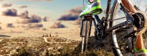 Dettaglio di un piede su un pedale di bicicletta, su terreno collinare arido con colline rocciose e cielo nuvoloso sullo sfondo.