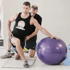 Uomo inginocchiato su un tappetino mentre esegue un esercizio con una palla da ginnastica viola, assistito da un altro uomo; scena ambientata in una palestra o studio di fisioterapia.
