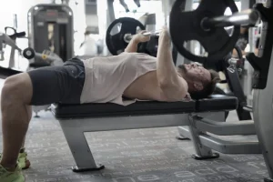 Uomo sdraiato su panca da allenamento mentre solleva un bilanciere con pesi in palestra, con indosso maglietta senza maniche e pantaloncini.