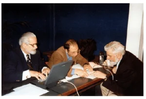 Tre uomini seduti a un tavolo in un ambiente professionale: due con giacche scure e uno con giacca marrone, tra cui Yuri Verkhoshansky con occhiali, in una discussione o analisi di documenti.