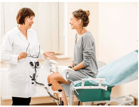 Una dottoressa in camice bianco parla con una paziente seduta su un lettino ginecologico in uno studio medico.