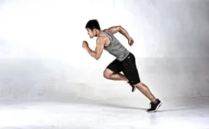 Uomo che corre indossando una canotta grigia, pantaloncini neri e scarpe da ginnastica, con uno sfondo semplice e chiaro.