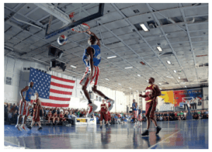Giocatore di basket in uniforme rossa, bianca e blu schiaccia il pallone nel canestro durante una partita in un palazzetto affollato.