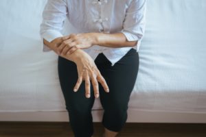 Persona seduta su un letto con camicia bianca e pantaloni scuri, che si tiene il polso per percepire i tremori, nel contesto di attività fisica adattata per soggetti parkinsoniani, durante un momento di recupero terapeutico