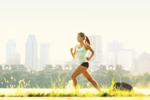 L'immagine mostra una donna che corre in un ambiente naturale con un prato verde in primo piano e una città con grattacieli sullo sfondo. La scena è illuminata dalla luce solare, creando un contrasto tra natura e contesto urbano. La donna indossa abbigliamento sportivo e si muove con energia in un’atmosfera serena.