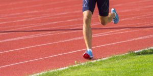 Nell'immagine si vede una persona che corre su una pista di atletica. Indossa pantaloncini aderenti blu e scarpe sportive azzurre. La pista è di colore rosso con linee bianche, e c'è anche dell'erba verde accanto alla pista.