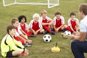 Bambini in divisa rossa ascoltano l’allenatore durante un allenamento di calcio giovanile, su un campo con porta e palloni.