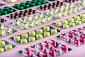 L'immagine mostra diverse confezioni di pillole di vari colori, disposte in modo ordinato su una superficie rosa. Le pillole sono di colore giallo, rosso, verde e marrone, rappresentando vari farmaci.