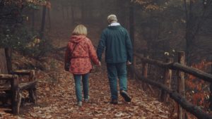 Due persone con giacche invernali, una rossa e una blu, che camminano mano nella mano lungo un sentiero autunnale nel bosco con foglie e leggera foschia.