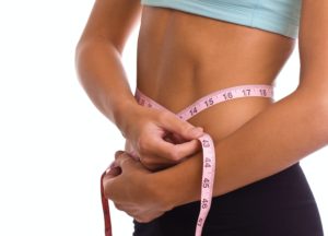 Persona con top azzurro e pantaloni neri misura la circonferenza vita con un nastro metrico rosa su sfondo bianco.