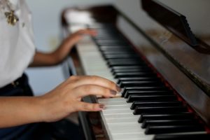 Mano che suona i tasti di un pianoforte, con maglietta bianca e collana dorata, in un’atmosfera tranquilla.