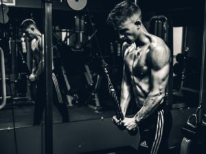 Uomo muscoloso a torso nudo che si allena in palestra con macchina a cavi, foto in bianco e nero con atmosfera drammatica.
