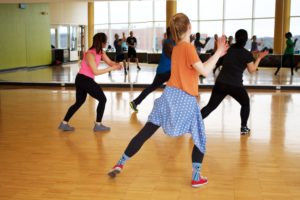 Gruppo di persone in abbigliamento sportivo che seguono una lezione di danza in una sala con pavimento in legno e pareti a specchio