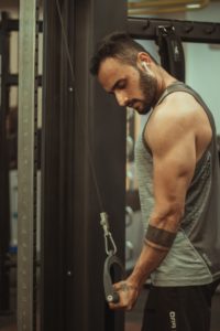 Uomo in palestra con canotta grigia e auricolari wireless che esegue esercizio tricipiti alla macchina per cavi; ambiente fitness con attrezzature.