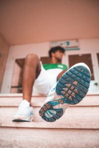 Dettaglio della suola sporca di una scarpa sportiva blu e nera, con persona seduta su gradini vista dal basso, pantaloni corti bianchi e maglietta verde e bianca, sfondo sfocato con porta.