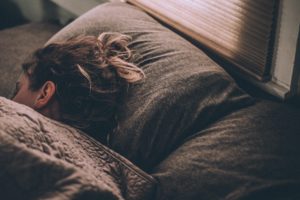 Persona distesa su un letto con cuscino grigio, coperta da una coperta, capelli raccolti e luce naturale che entra dalla finestra.