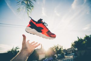 Scarpa sportiva arancione con dettagli neri e suola bianca sospesa in aria, lanciata da una mano, con cielo azzurro e nuvole sullo sfondo