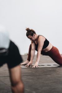 Donna che esegue esercizi di stretching o yoga su un tappetino, con top nero e leggings marroni, in uno studio o palestra; figura sfocata in primo piano.