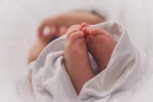 Piedini di un neonato avvolti in un panno bianco, al centro dell’immagine con sfondo sfocato che suggerisce il corpo del bambino sdraiato.