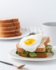 Sandwich con pane integrale, insalata e uovo fritto su piatto bianco, con piatto di fette di pane e tazza sullo sfondo