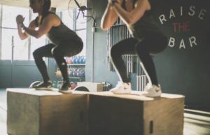 Due persone eseguono salti su box in legno durante un allenamento funzionale in palestra, con attrezzi e la scritta "RAISE THE BAR" sullo sfondo.