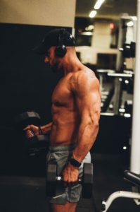 Uomo muscoloso che si allena con i pesi in palestra, indossando cappellino nero, cuffie e pantaloncini grigi, circondato da attrezzature fitness.