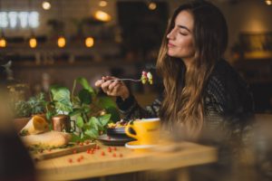 Donna che gusta un pasto con gli occhi chiusi, seduta a un tavolo con ingredienti freschi e una ciotola di cibo.