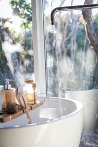 Vasca da bagno bianca davanti a una finestra con vista sugli alberi, con acqua corrente e vassoio in legno con accessori da bagno come sapone, spugna e pennello.