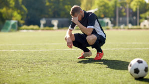 L'immagine mostra un calciatore accovacciato su un campo, con una mano sul viso in segno di frustrazione. Il pallone vicino e l’ambiente naturale suggeriscono un contesto reale di partita o allenamento. La scena evidenzia il lato emotivo del calcio e l’importanza della psicologia dello sport nei momenti di pressione o insuccesso.