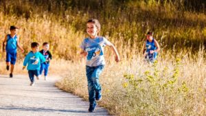 L’immagine mostra un bambino che corre felice lungo un sentiero naturale, seguito da altri. Con indosso jeans e maglietta grigia, è immerso in un ambiente soleggiato e verde. La scena trasmette energia, libertà e benessere, evidenziando l’importanza del movimento e del gioco all’aperto per la crescita sana dei bambini.