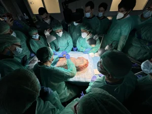 Personale medico e studenti con camici e mascherine osservano una dissezione anatomica attorno a un tavolo operatorio illuminato.