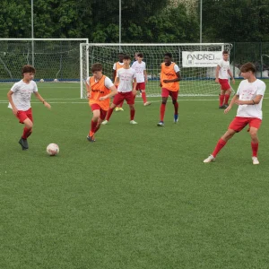 Giovani calciatori durante un allenamento su campo sintetico, con maglie bianche e pantaloncini rossi, impegnati in un’azione di gioco.