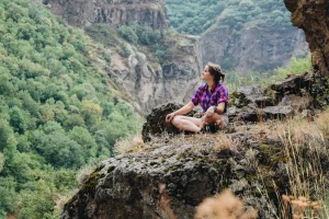 Persona seduta su una grande roccia in montagna, con camicia a quadri e pantaloncini, che osserva il paesaggio naturale circostante.