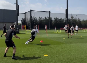 Atleta su campo in erba sintetica che esegue un cambio di direzione con palla medica, circondato da coni e attrezzatura per agilità durante un allenamento sportivo.