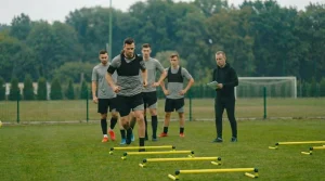 Uomo che corre su ostacoli bassi durante un allenamento di agilità, osservato da compagni e allenatore su un campo d’erba.