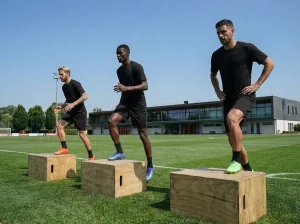 Tre atleti eseguono esercizi di pliometria su plyo box in un campo sportivo, indossando abbigliamento nero e scarpe da calcio colorate.